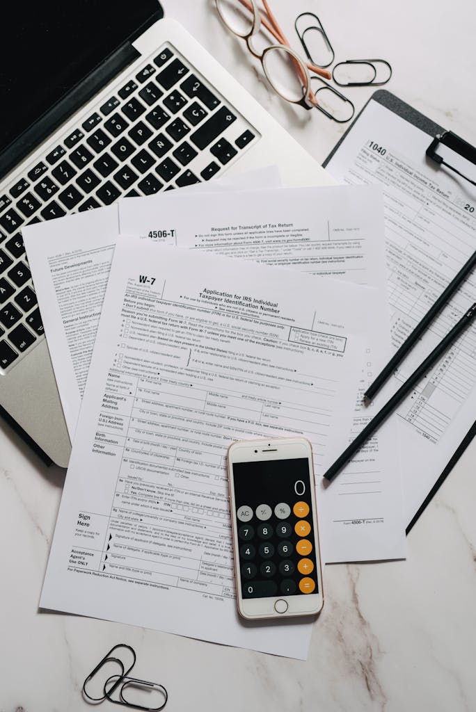 A desk with W-7 tax forms, a calculator, and a laptop for tax preparation.